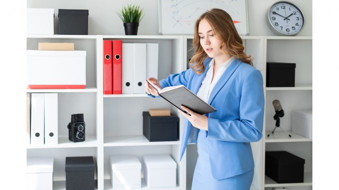 A beautiful young girl stands near a rack in the office and holds an open book in her hands.